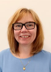 Malin mot neutral bakgrund. Hon har ljusblå tröja och halsband i guld med hjärtformat hänge. Hon ler mot kameran.